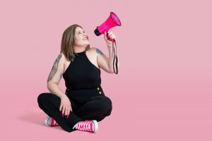 Shh! PR founder Amie-Leigh sat on the floor against a pink backdrop with her left arm in the air holding a pink megaphone